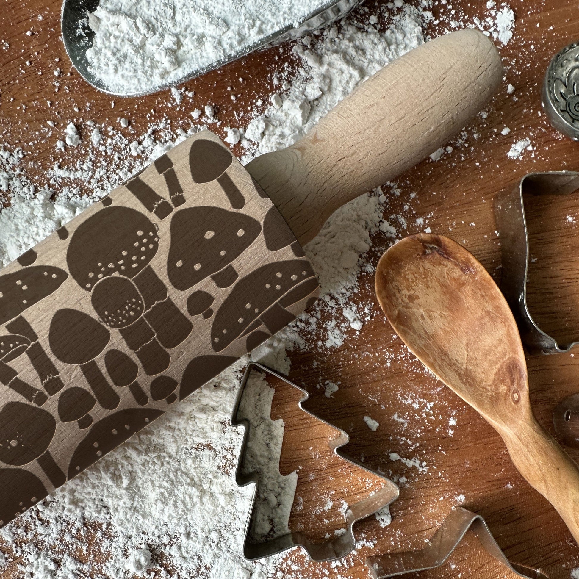 Rolling pin with mushroom pattern on a wooden surface with flour and cookie cutters.