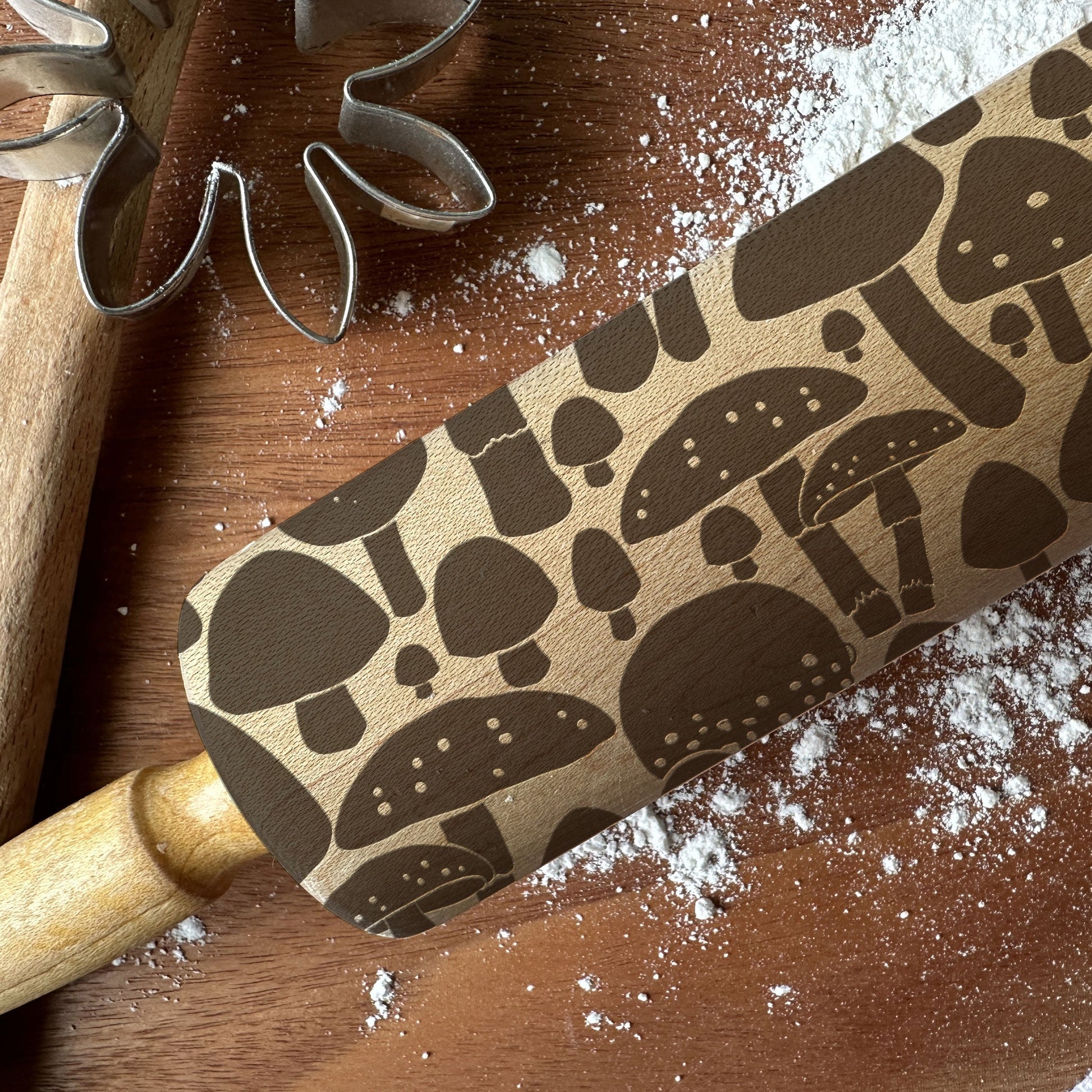 Rolling pin with mushroom pattern on a wooden surface with flour and cookie cutters.