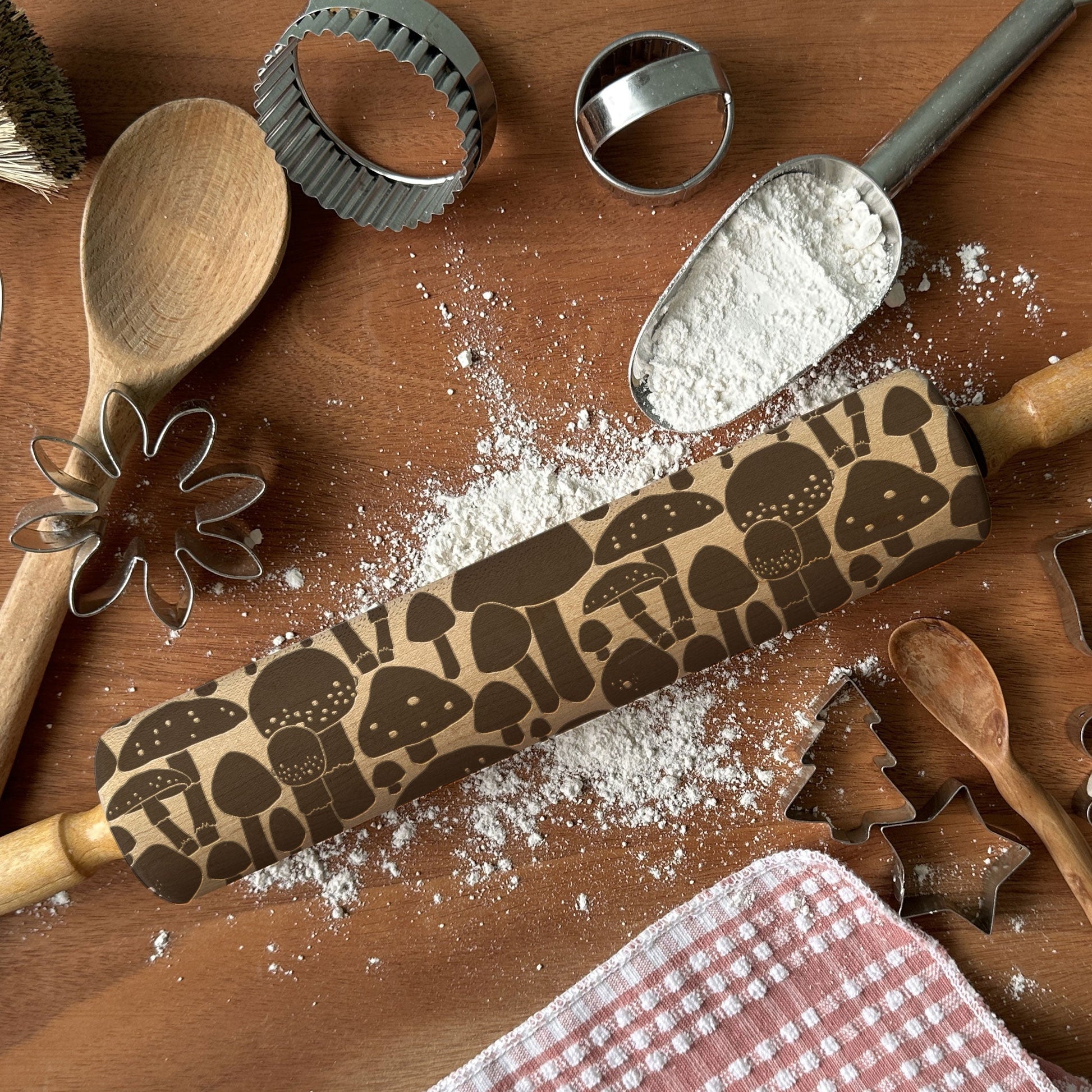 Baking tools including a rolling pin with mushroom pattern, wooden spoon, and flour on a wooden surface.