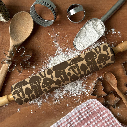 Baking tools including a rolling pin with mushroom pattern, wooden spoon, and flour on a wooden surface.