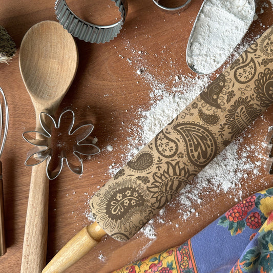 Baking tools including a wooden spoon, rolling pin with floral patterns, and cookie cutters on a flour-dusted surface.
