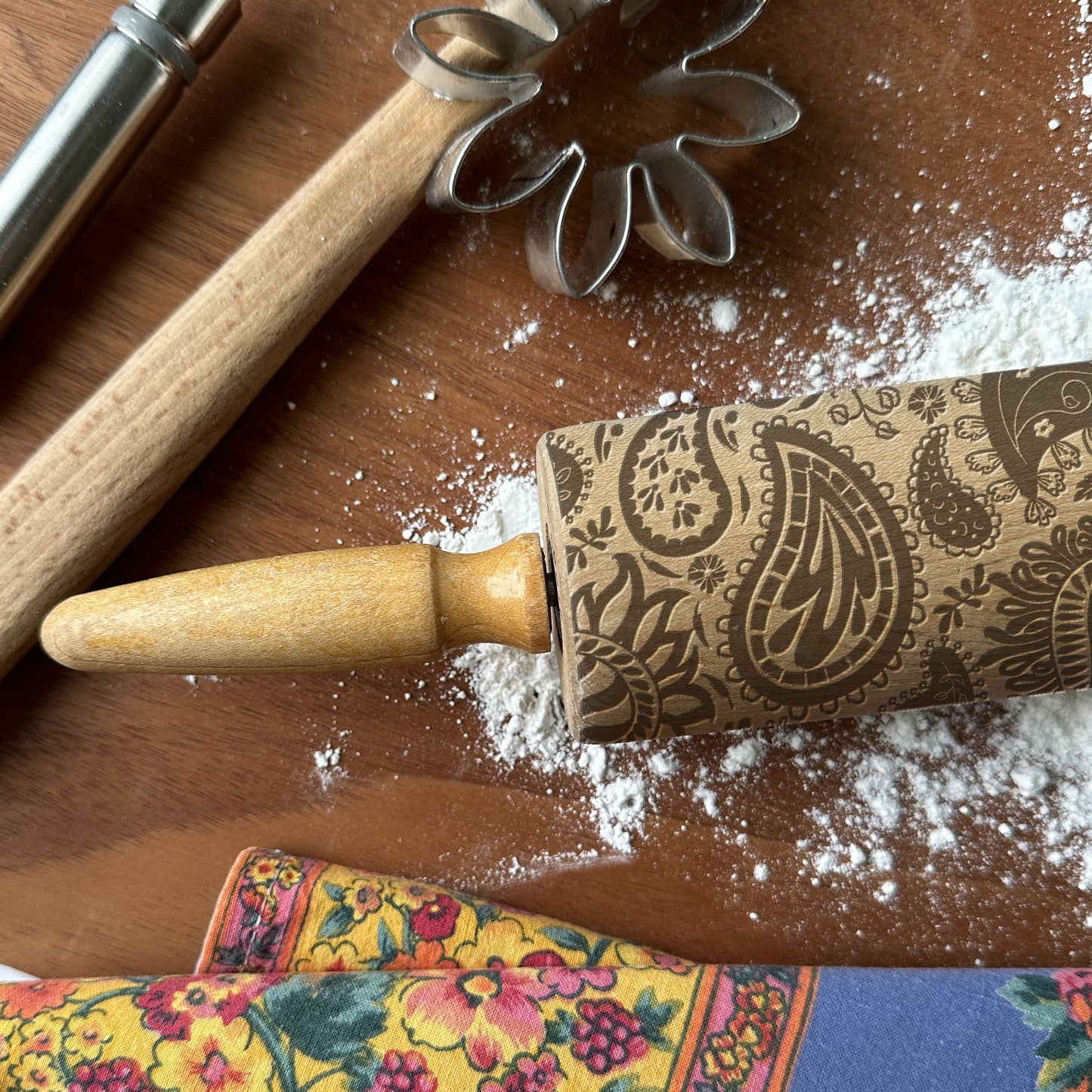 Decorative rolling pin with paisley pattern on a wooden surface with flour and cookie cutters.