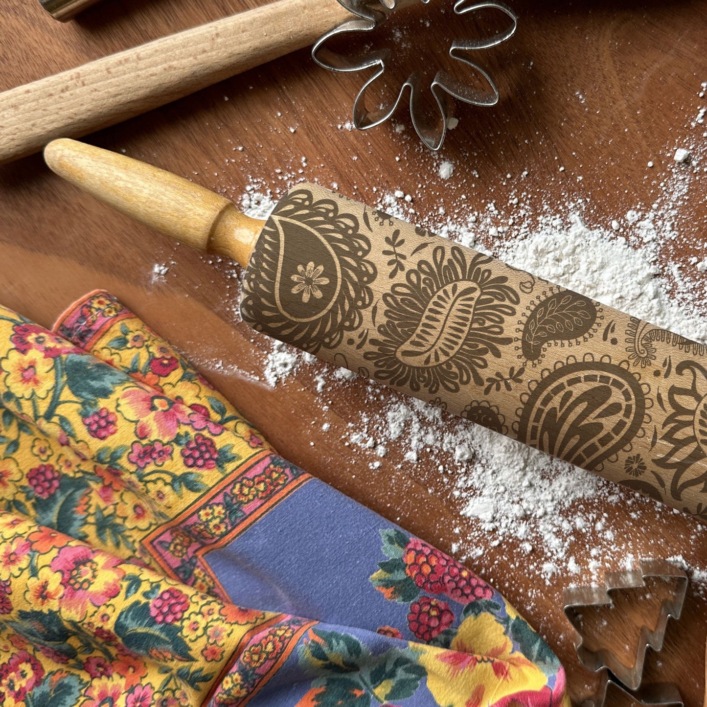 Decorative rolling pin with floral patterns on a wooden surface with flour and cookie cutters.