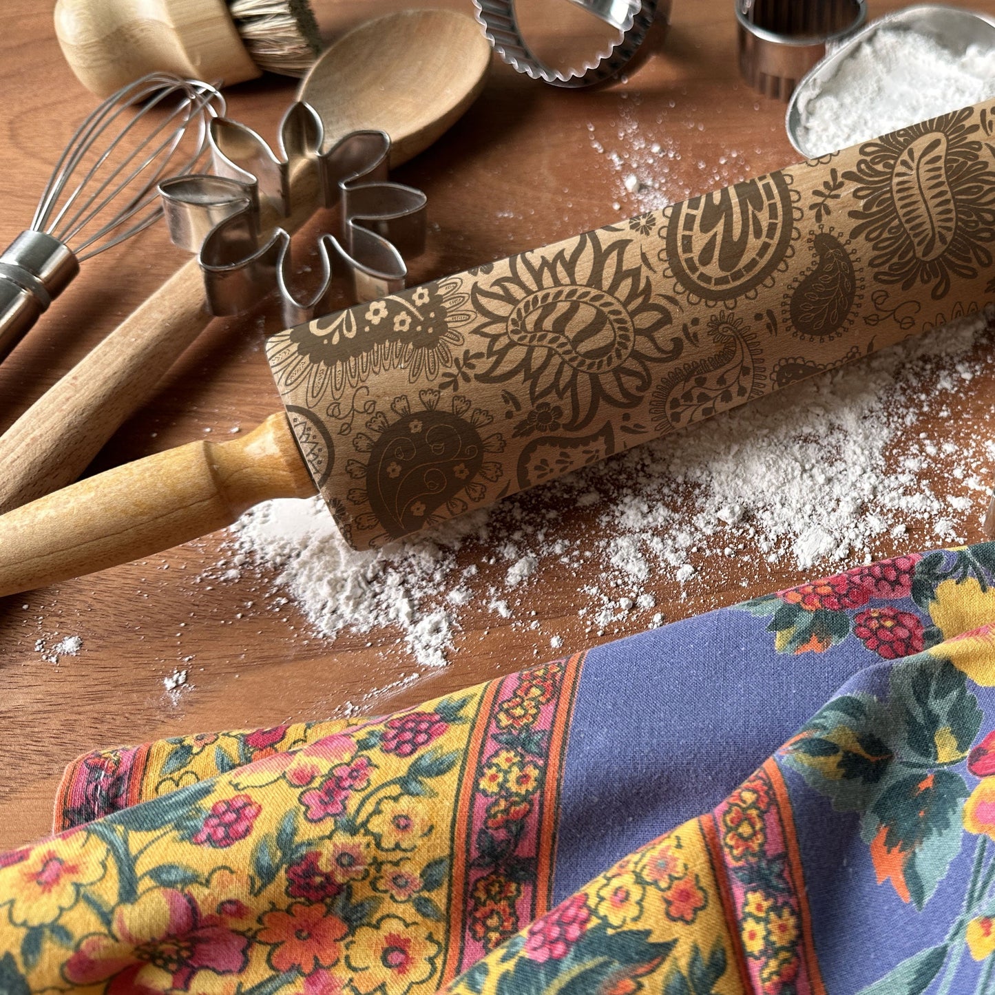 Decorative rolling pin with floral patterns on a wooden surface with kitchen utensils and a colorful towel.
