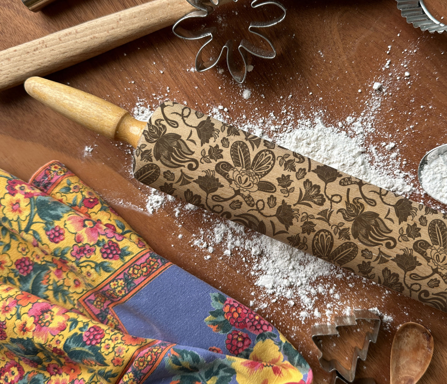 Decorative rolling pin with floral patterns on a wooden surface with flour and a colorful cloth.
