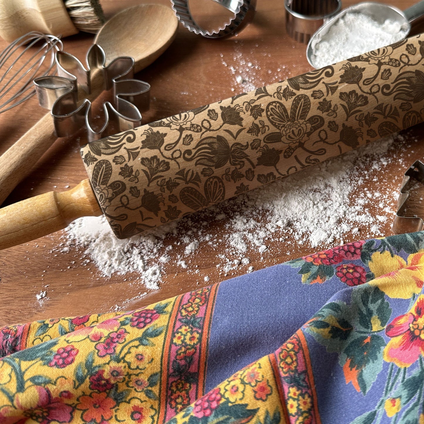 Decorative rolling pin with floral pattern on a wooden surface with flour and a colorful kitchen towel.