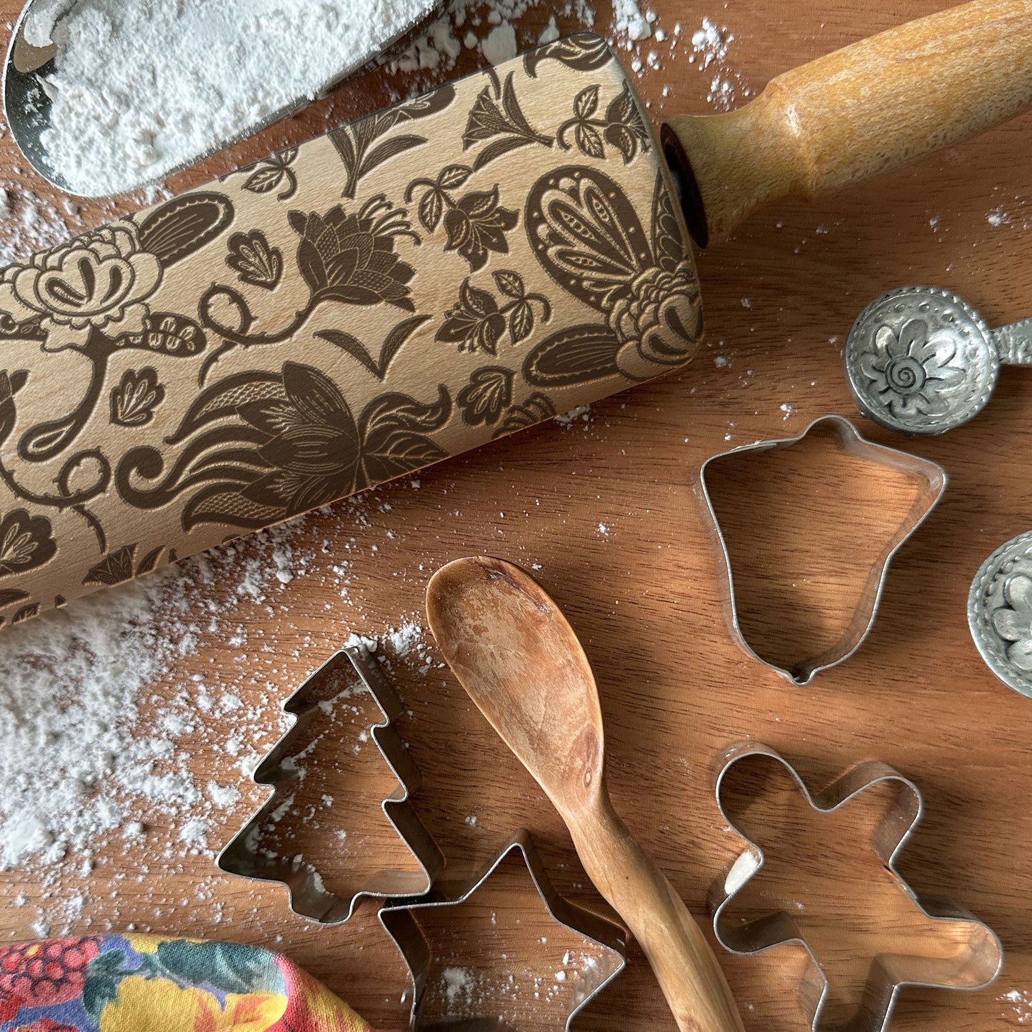 Baking tools including a floral-patterned rolling pin, cookie cutters, and measuring spoons on a wooden surface with flour.