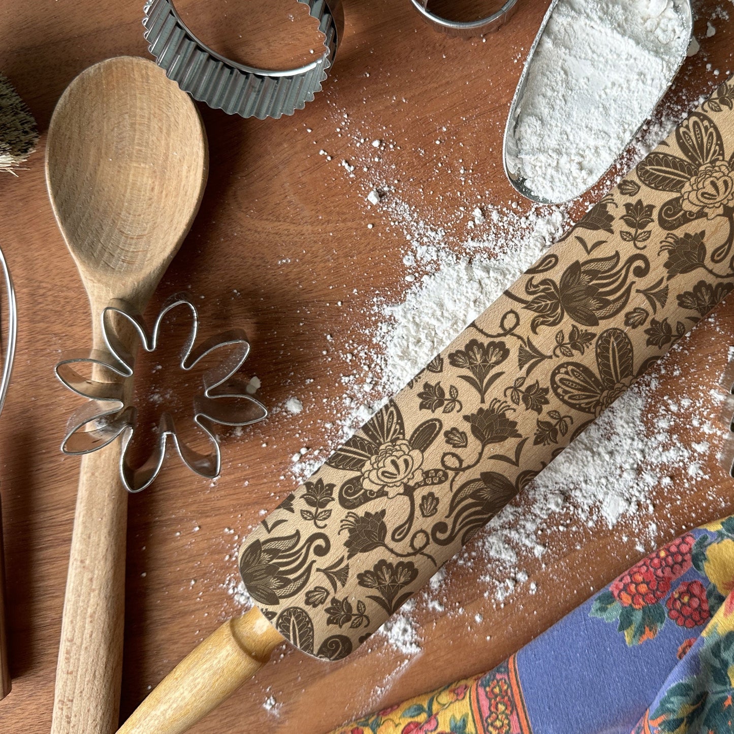 Baking tools including a wooden spoon, rolling pin with floral patterns, and cookie cutters on a wooden surface.