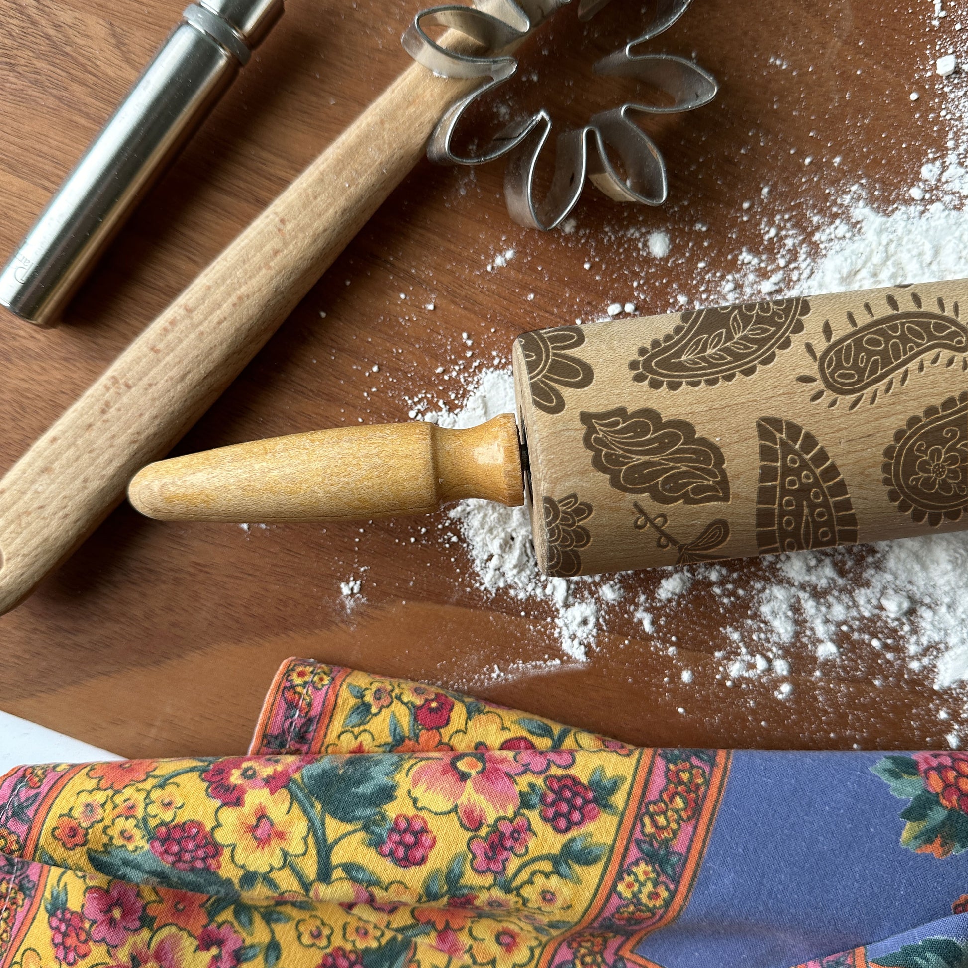 Decorative rolling pin with paisley patterns on a wooden surface with flour and cookie cutters.