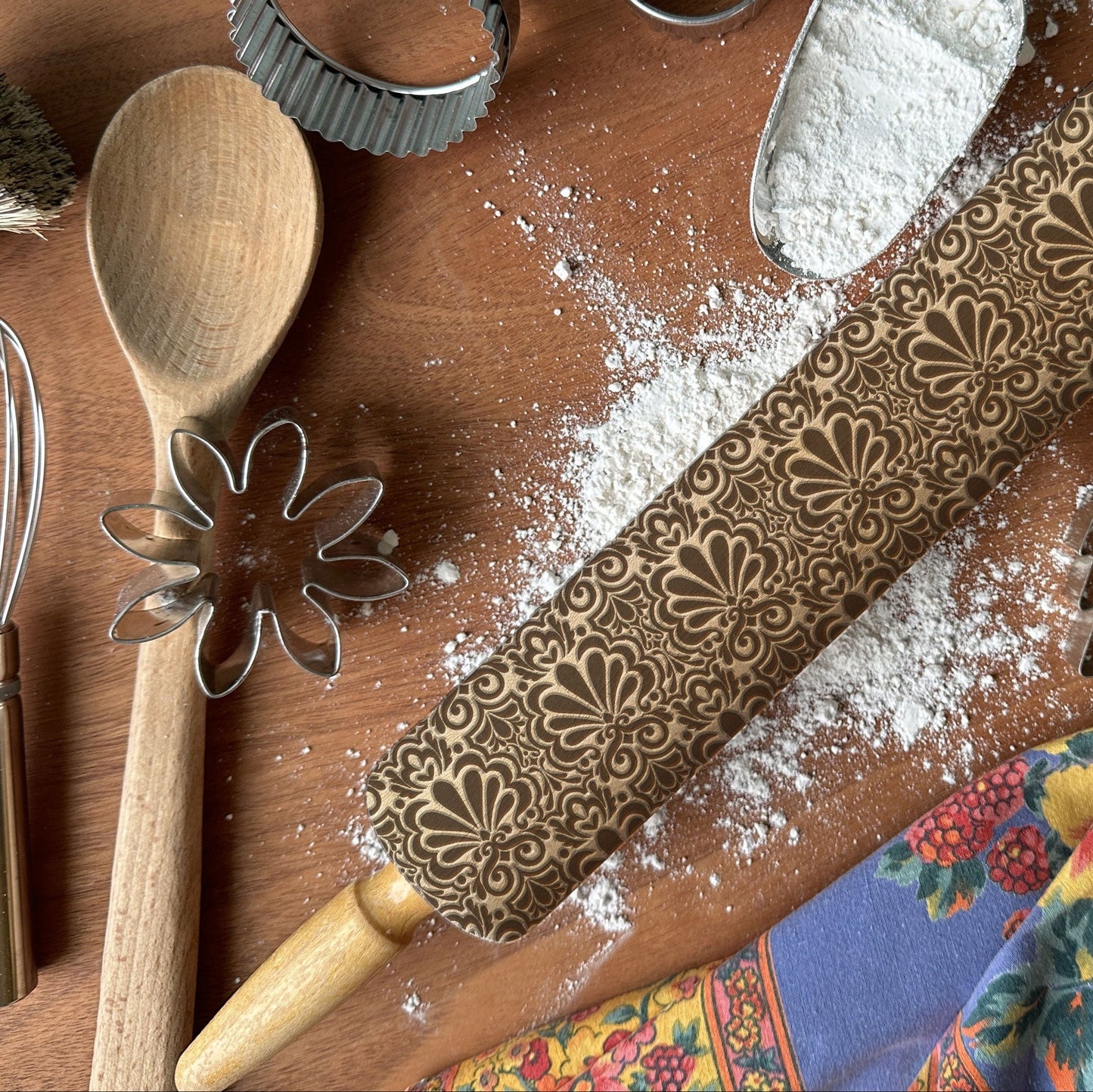 Baking tools including a wooden spoon, rolling pin with floral patterns, and flour on a wooden surface.
