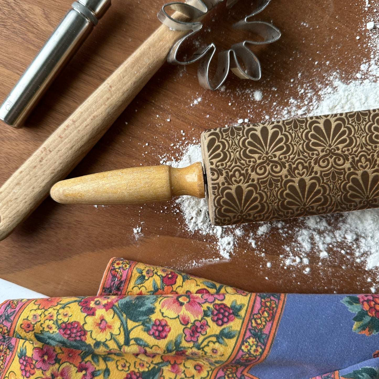 Decorative rolling pin with floral pattern on a wooden surface with flour and cookie cutters.