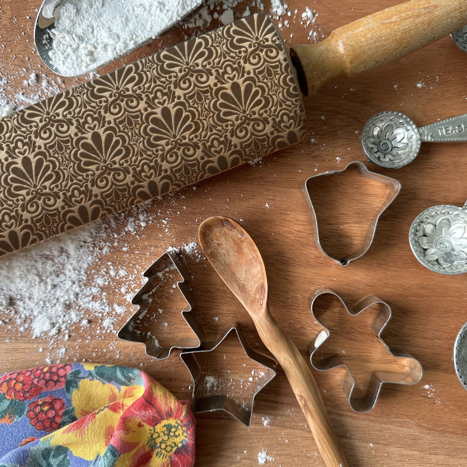 Baking tools including a patterned rolling pin, cookie cutters, and measuring spoons on a wooden surface with flour.