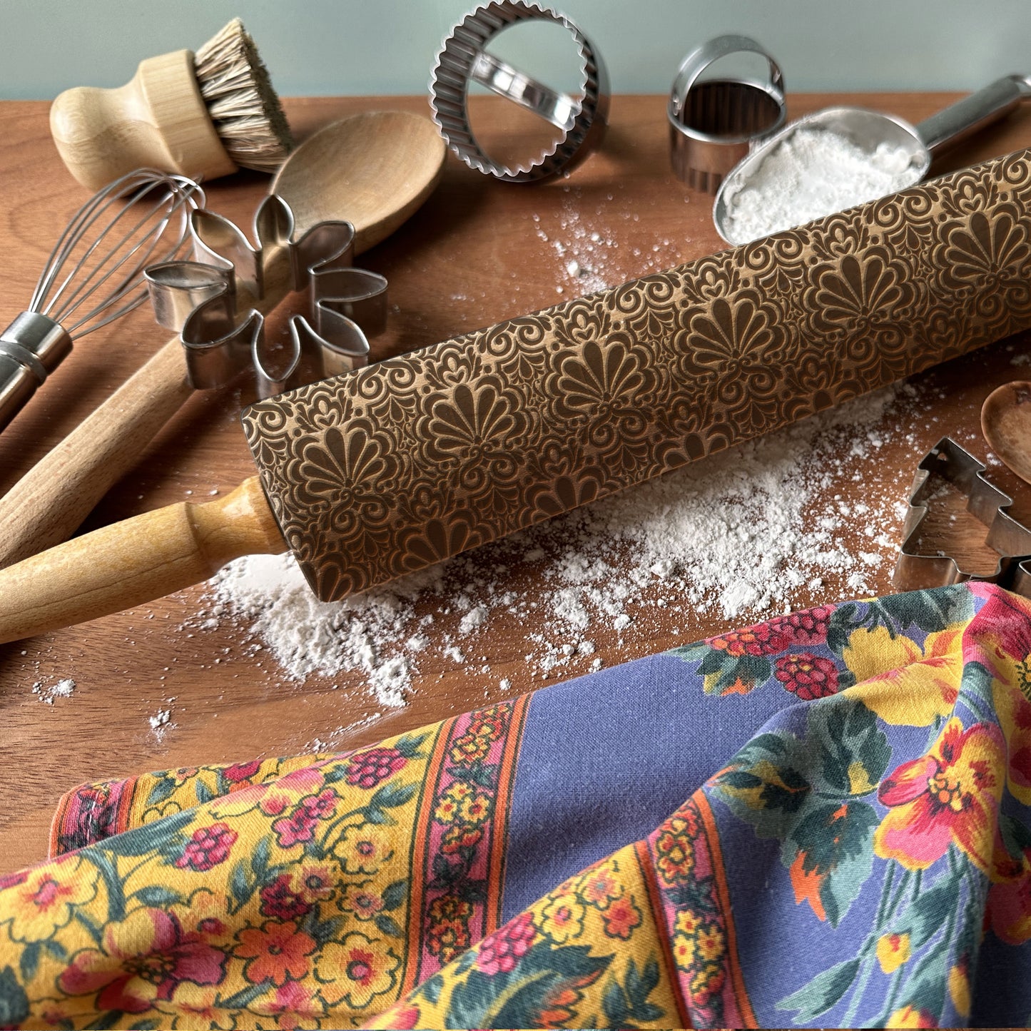 Decorative rolling pin with embossed pattern on a wooden surface with kitchen utensils and a colorful towel.