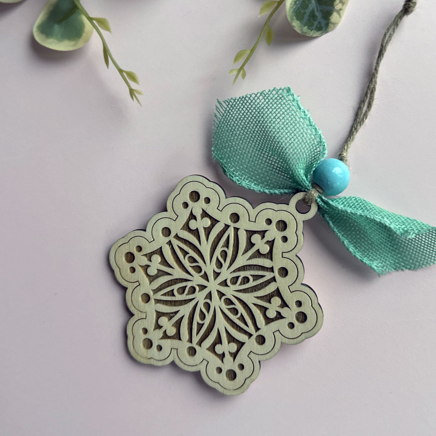 Decorative snowflake ornament with teal ribbon and blue bead on a light pink background