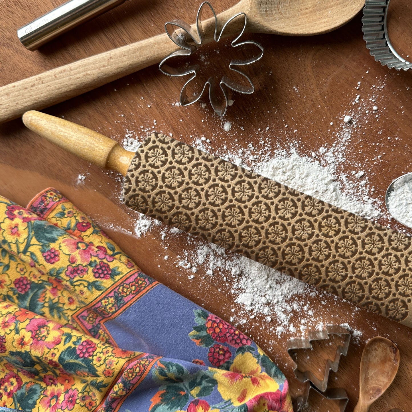 Decorative rolling pin with floral pattern on a wooden surface with flour and cookie cutters.