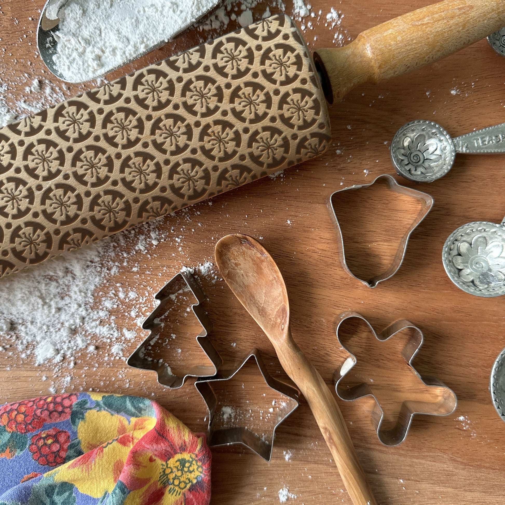 Baking tools including a patterned rolling pin, cookie cutters, and measuring spoons on a wooden surface with flour.