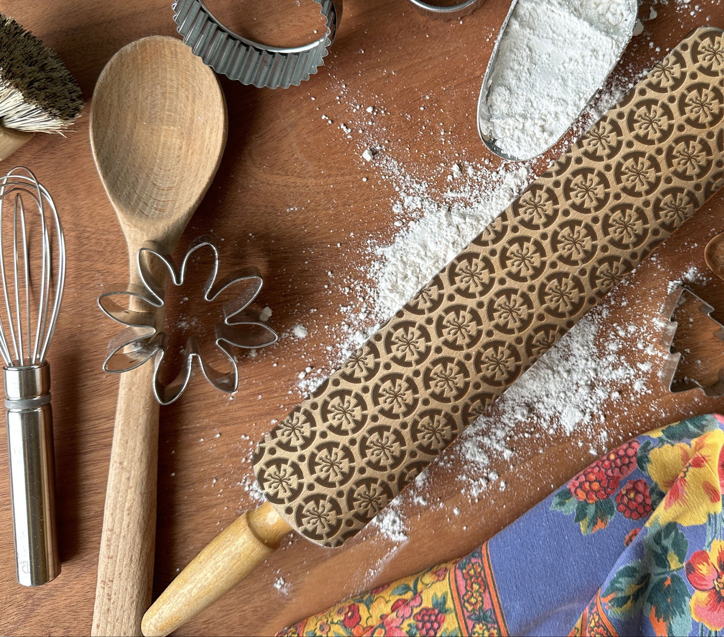 Baking tools including a rolling pin with floral patterns, wooden spoon, whisk, and cookie cutters on a wooden surface with flour.