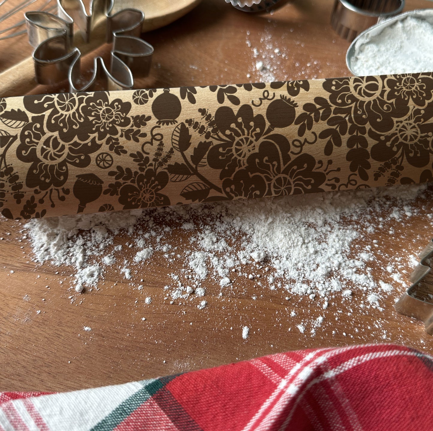 Decorative rolling pin with floral pattern on a wooden surface with flour and cookie cutters.
