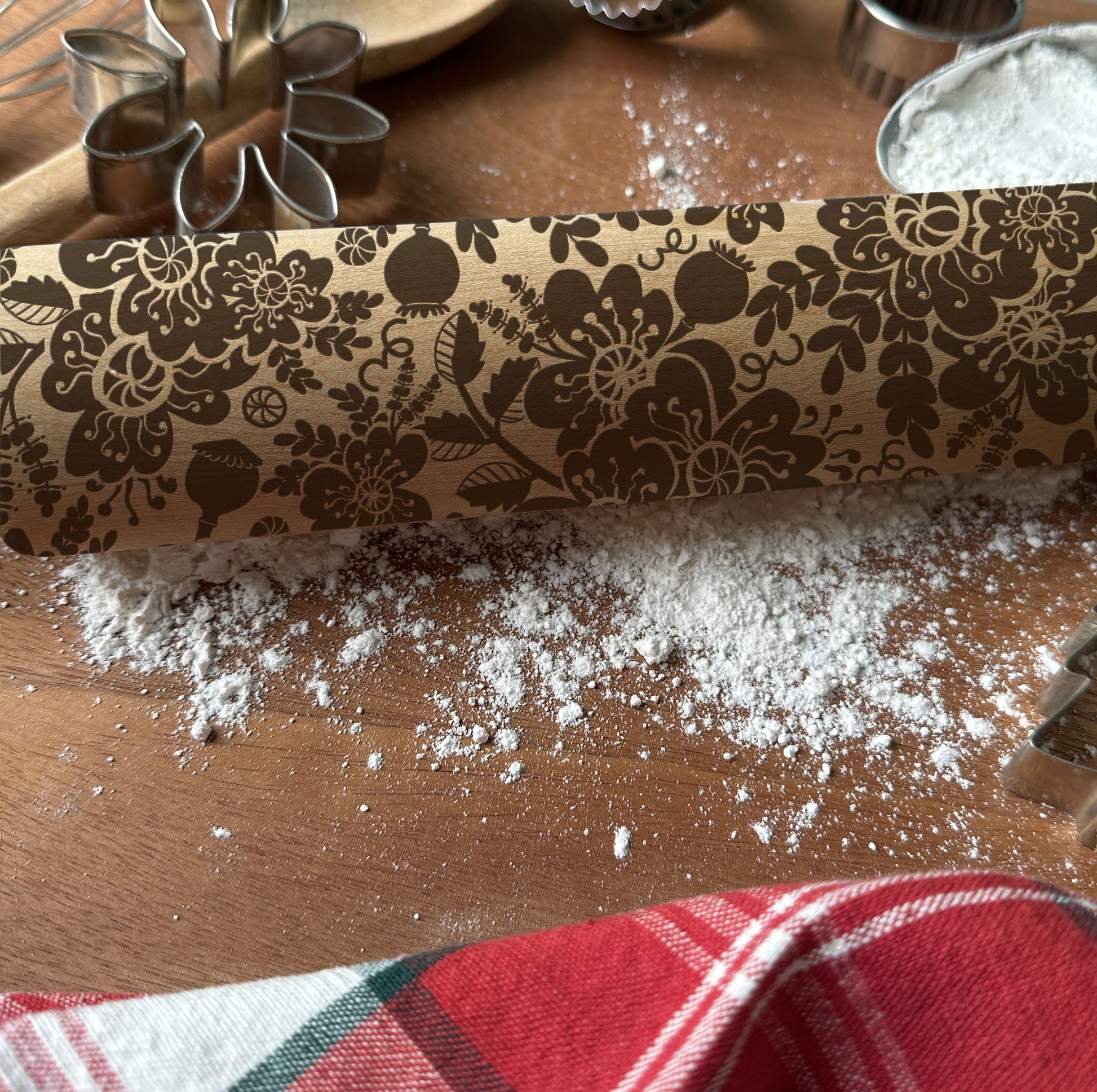 Decorative rolling pin with floral pattern on a wooden surface with flour and cookie cutters.