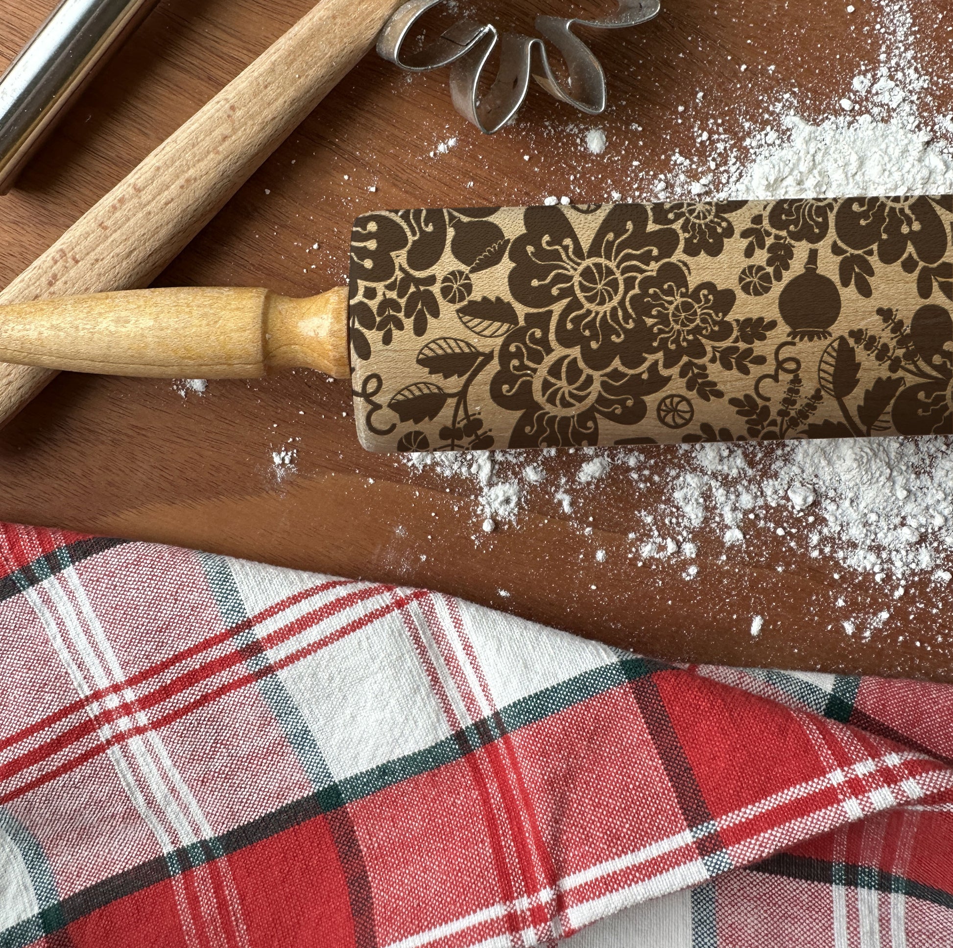 Decorative rolling pin with floral pattern on a wooden surface with flour and plaid fabric.