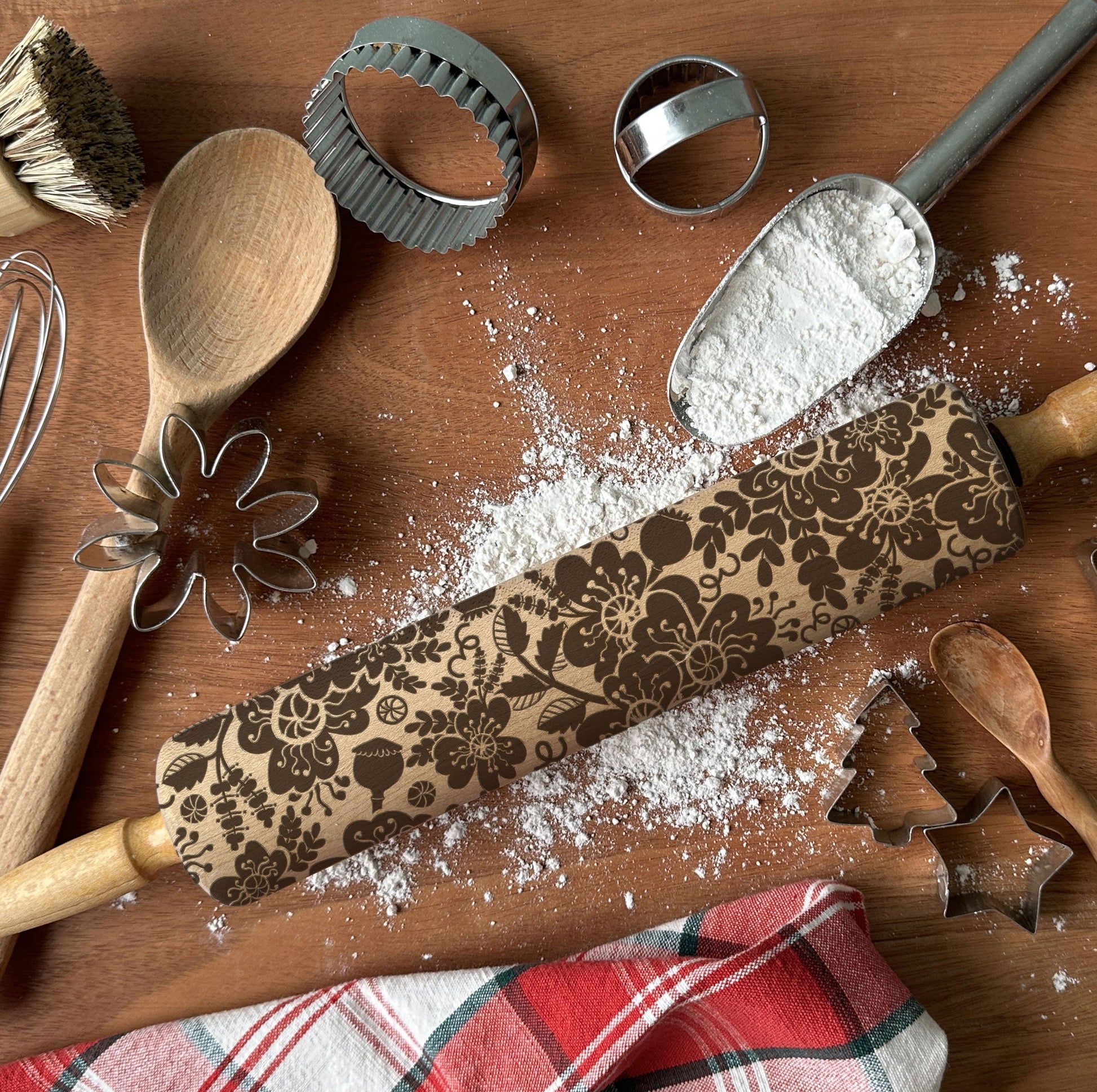 Baking tools including a rolling pin with a decorative pattern, wooden spoon, whisk, and cookie cutters on a wooden surface.