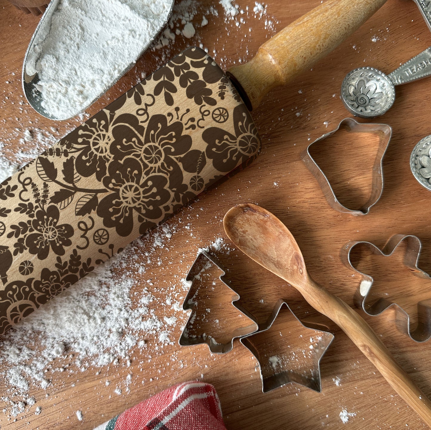 Wooden rolling pin with floral pattern, metal cookie cutters, and wooden spoon on a wooden surface with flour.
