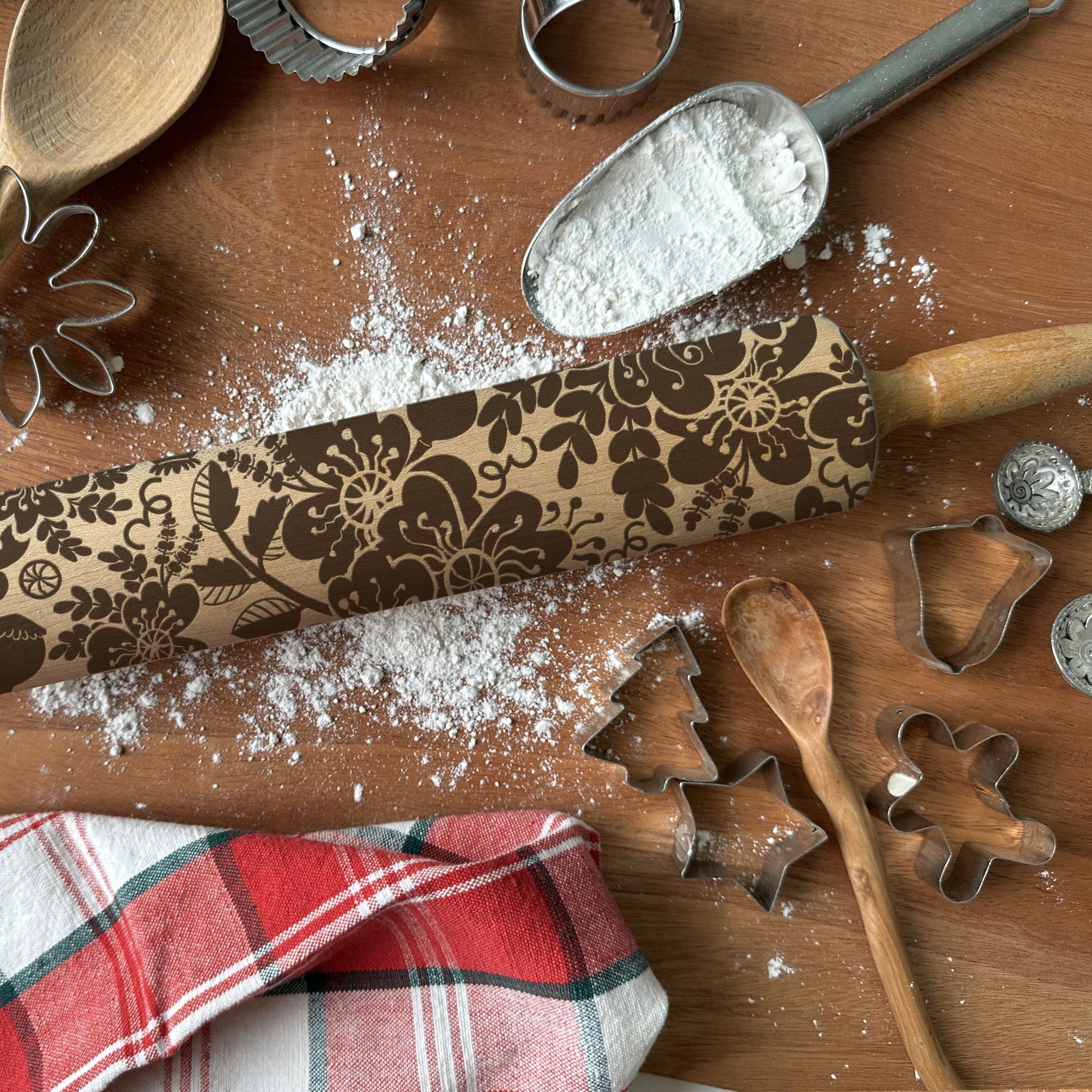 Decorative rolling pin with floral patterns on a wooden surface with flour, cookie cutters, and a plaid cloth.