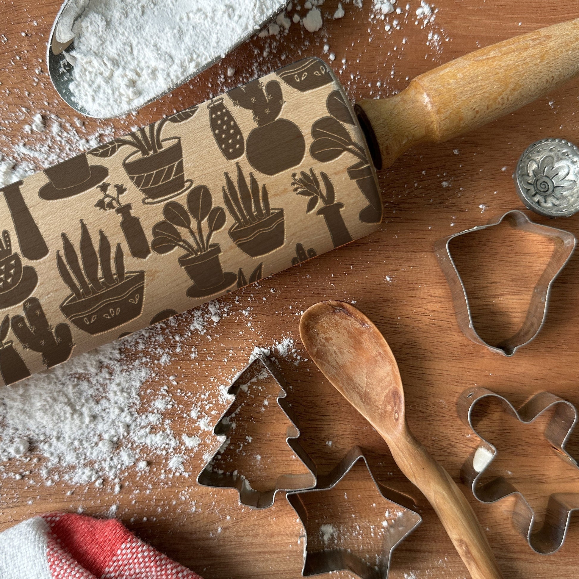 Rolling pin with plant pattern, cookie cutters, and wooden spoon on a wooden surface with flour.