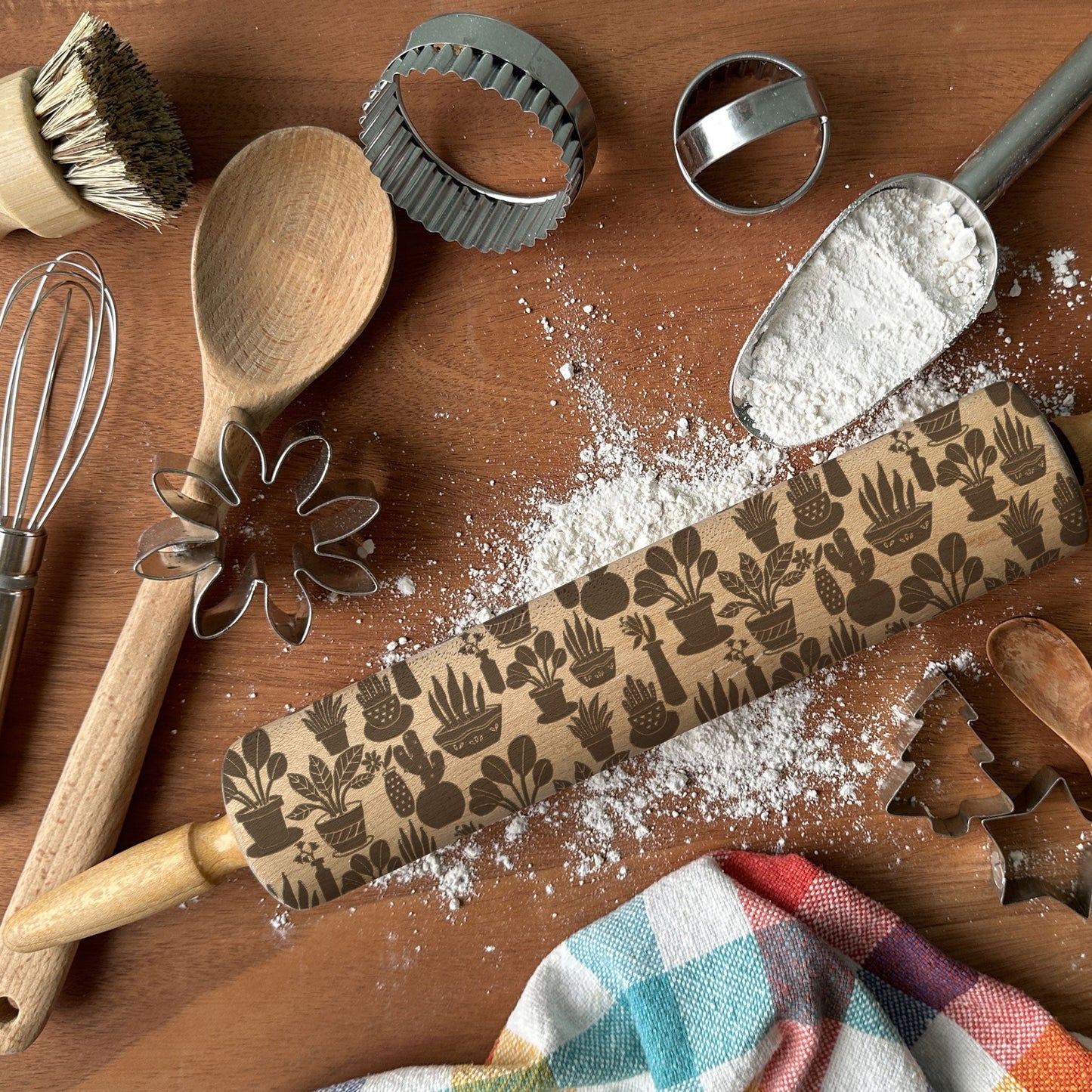 Baking tools including a rolling pin with a pattern, wooden spoon, whisk, and cookie cutters on a wooden surface with flour.
