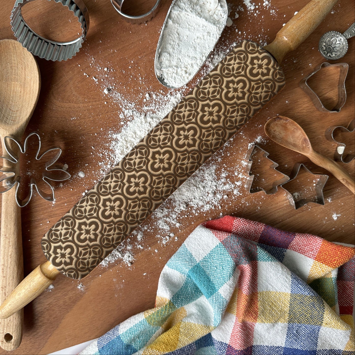 Baking tools including a wooden spoon, rolling pin with patterns, and cookie cutters on a wooden surface with flour.