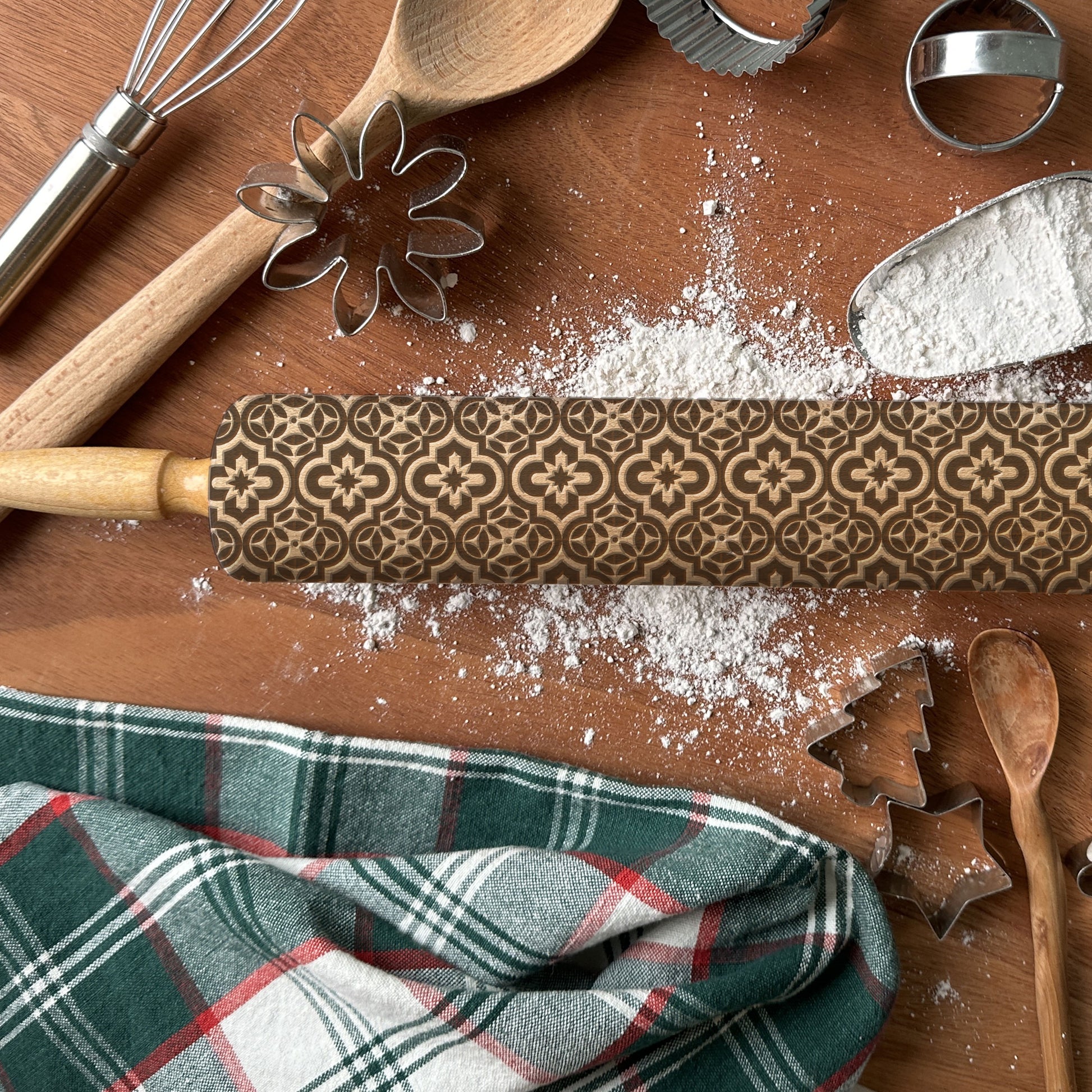 Patterned rolling pin on a wooden surface with flour, cookie cutters, and a plaid towel.