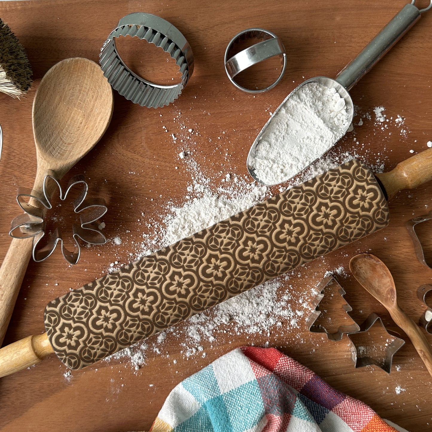 Wooden rolling pin with embossed pattern, flour, wooden spoon, and cookie cutters on a wooden surface.