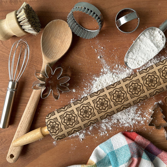 Baking tools including a rolling pin with embossed designs, wooden spoon, whisk, and cookie cutters on a wooden surface.