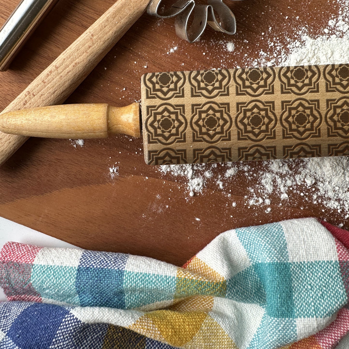 Decorative rolling pin with geometric patterns on a wooden surface with flour and a checkered cloth.