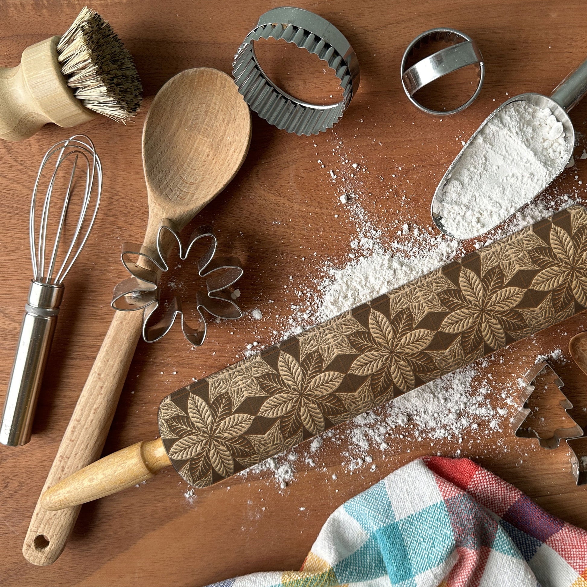 Baking tools including a rolling pin with floral patterns, wooden spoon, whisk, and cookie cutters on a wooden surface.