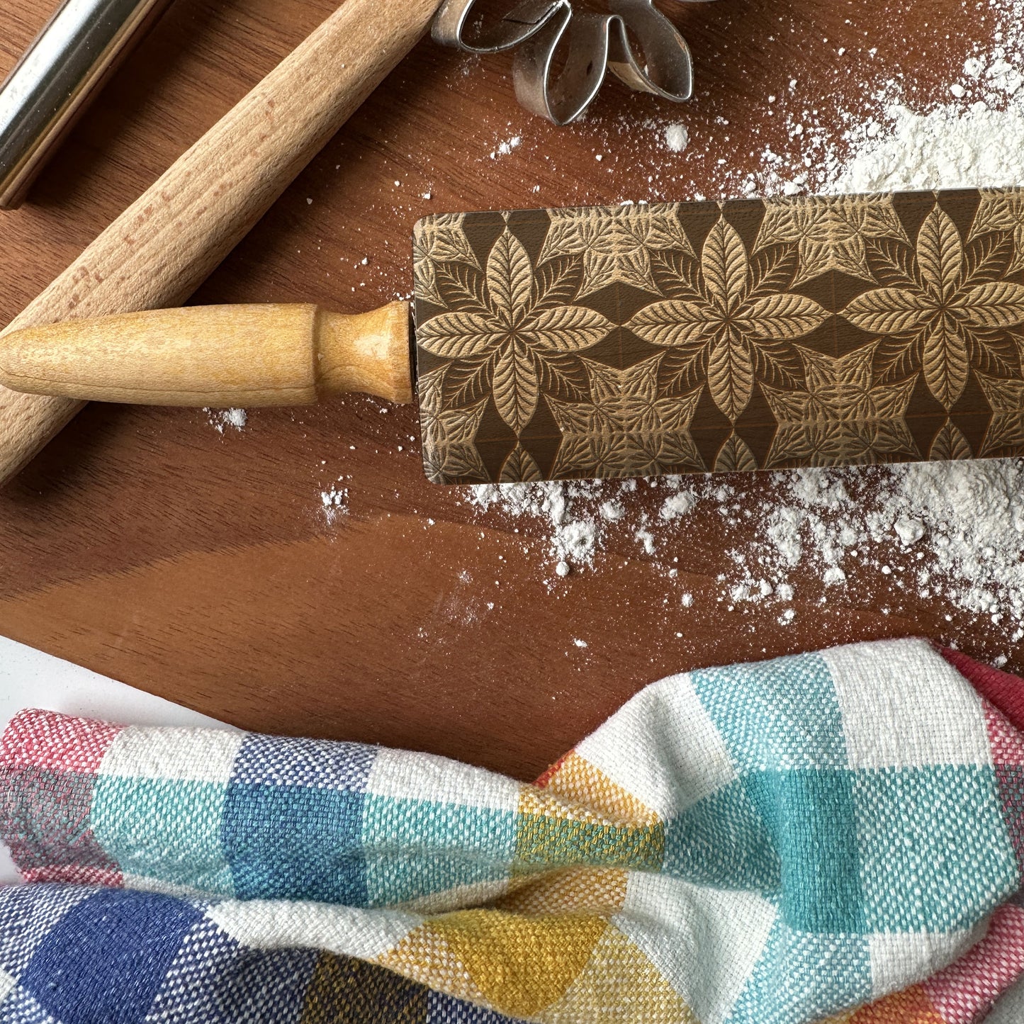 Decorative rolling pin with floral pattern on a wooden surface with flour and a checkered cloth.