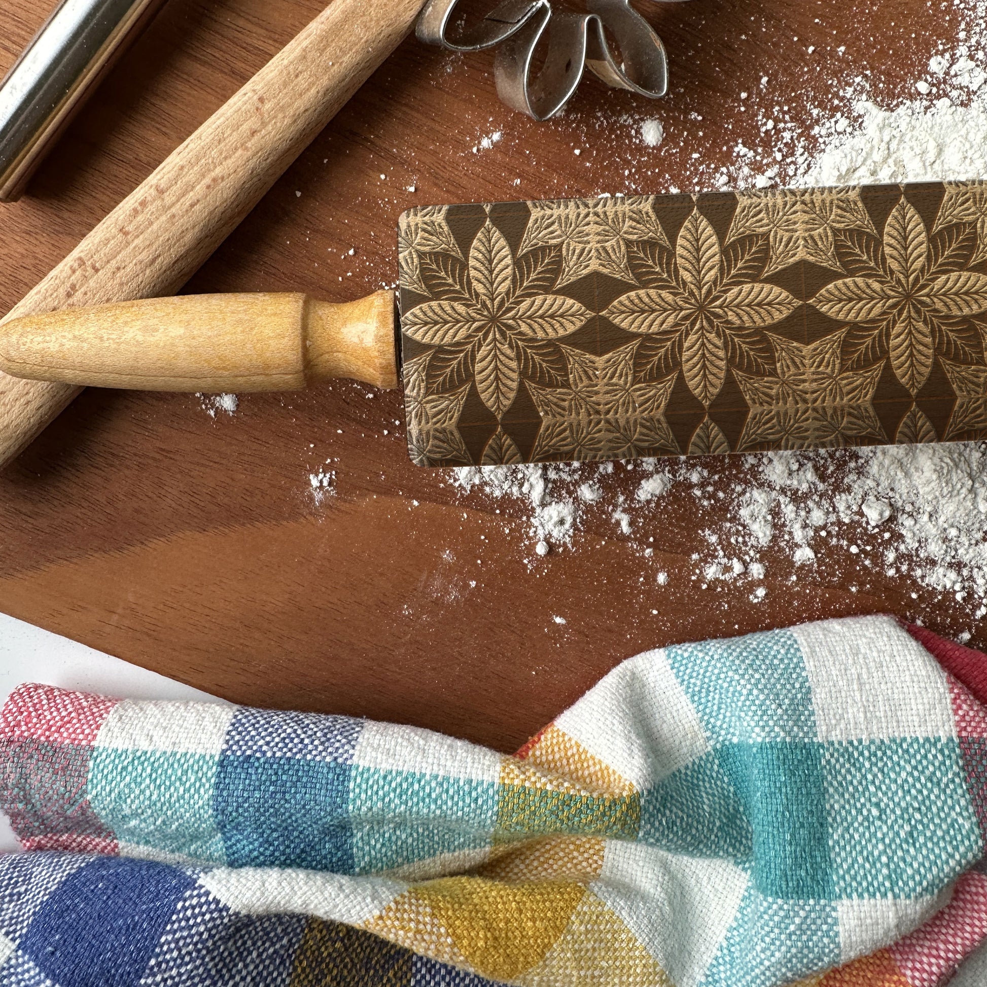 Decorative rolling pin with floral pattern on a wooden surface with flour and a checkered cloth.