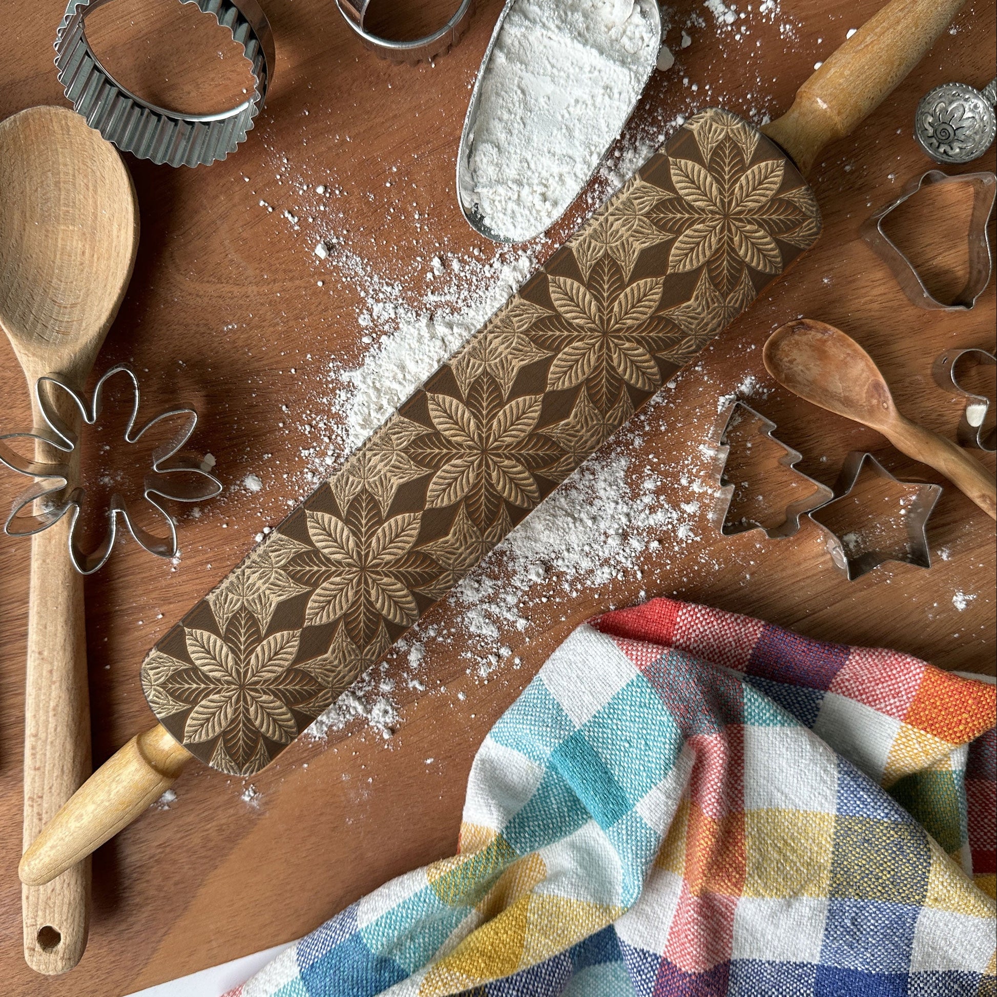 Baking tools including a rolling pin with a star pattern, wooden spoon, whisk, and cookie cutters on a wooden surface with flour.