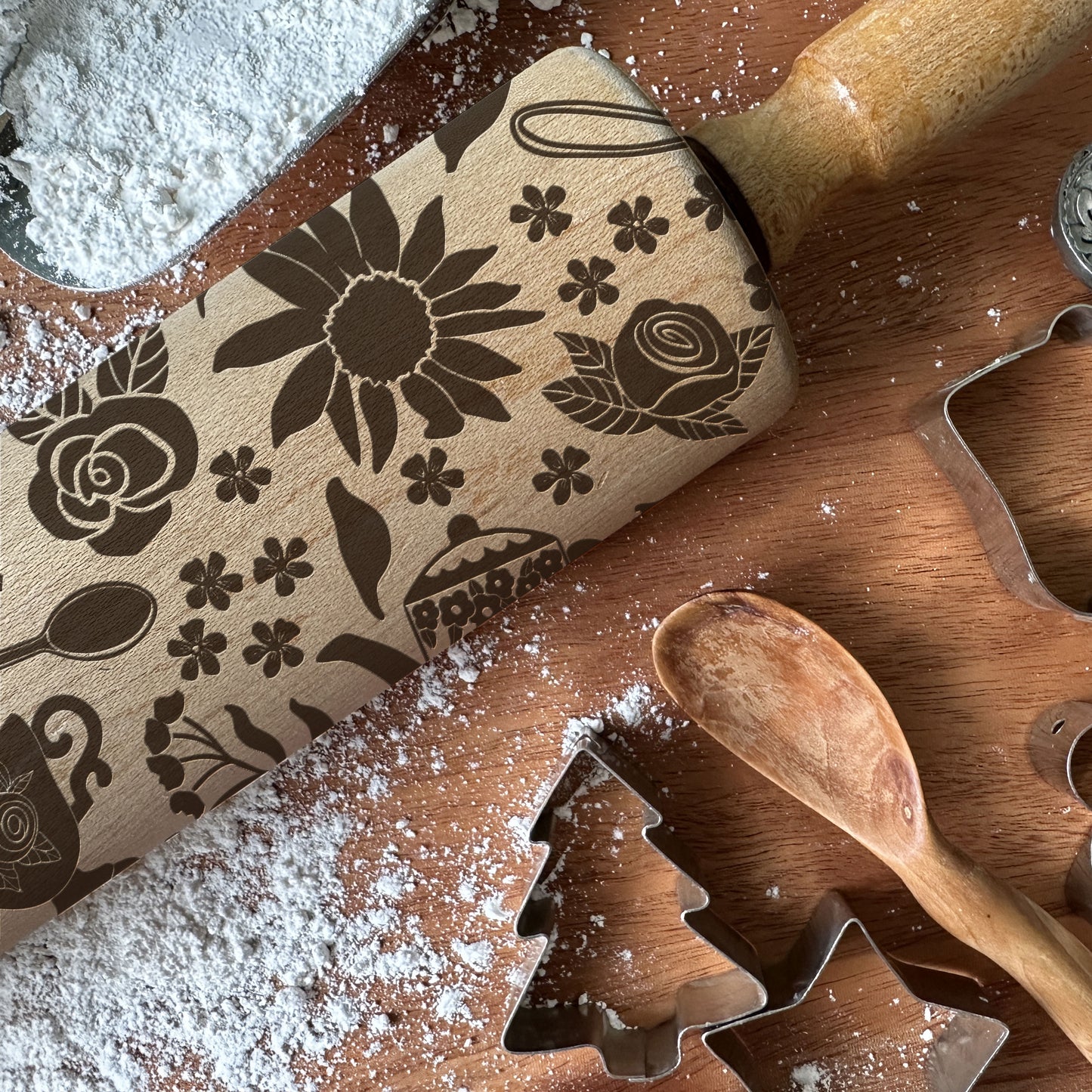 Wooden rolling pin with floral designs on a wooden surface with cookie cutters and flour.