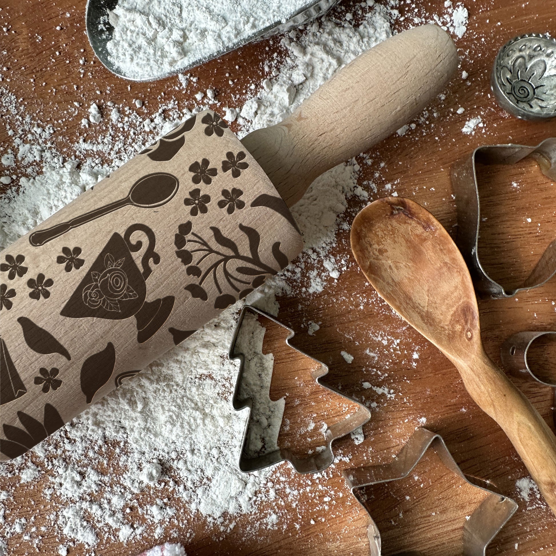 Wooden rolling pin with embossed designs on a flour-dusted surface with cookie cutters and a wooden spoon.