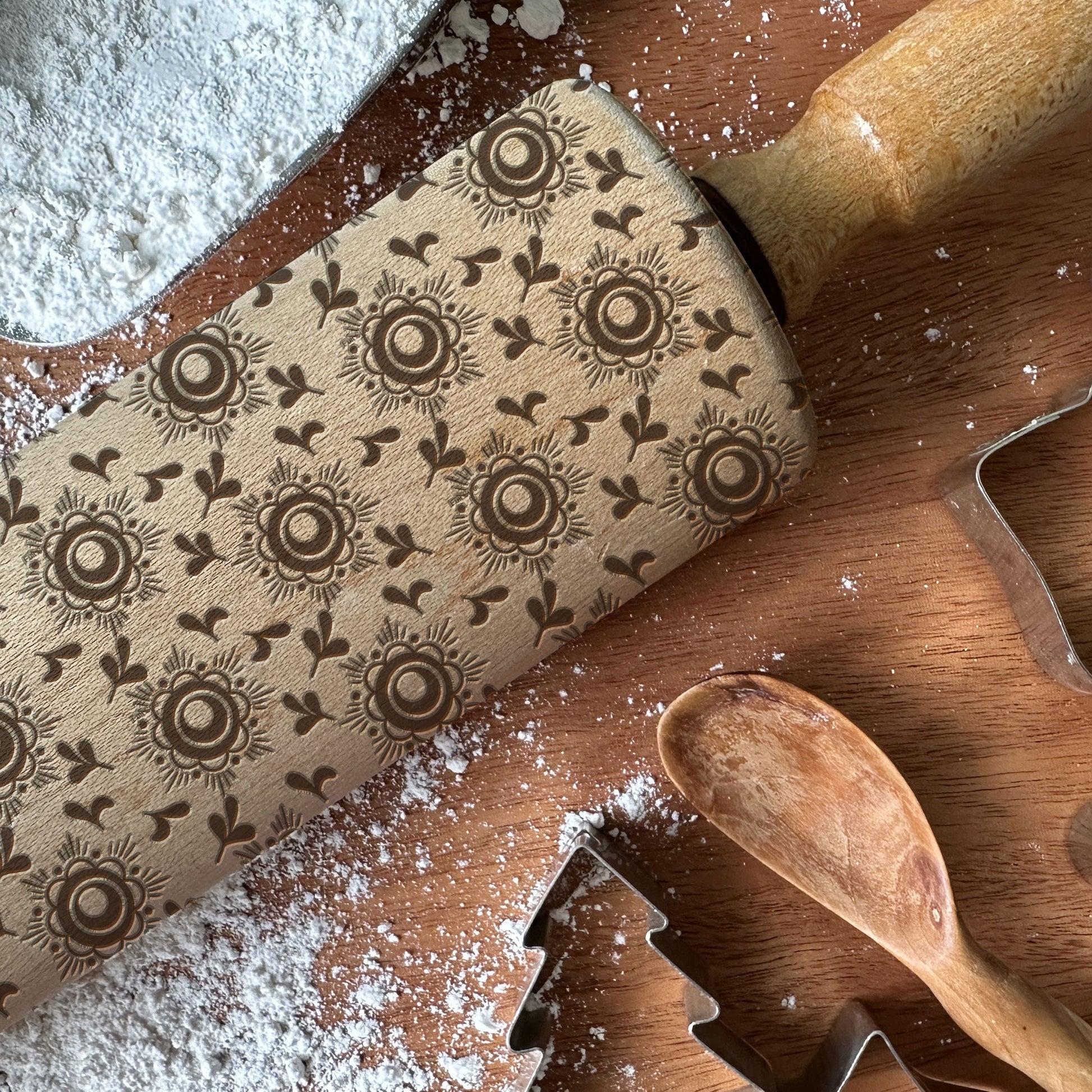 Decorative rolling pin with floral patterns on a wooden surface with flour and cookie cutters.