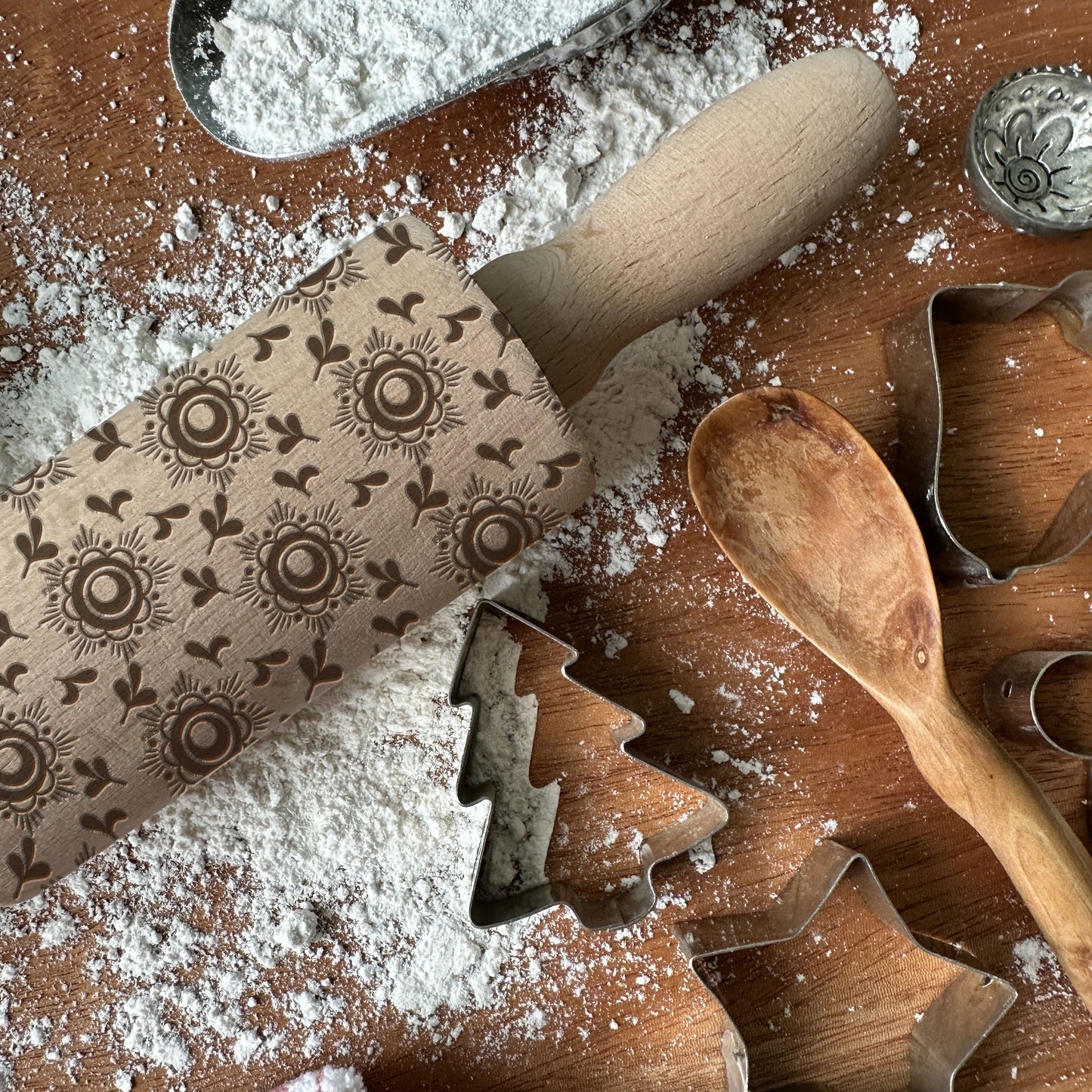 Wooden rolling pin with floral patterns on a wooden surface with flour and cookie cutters.