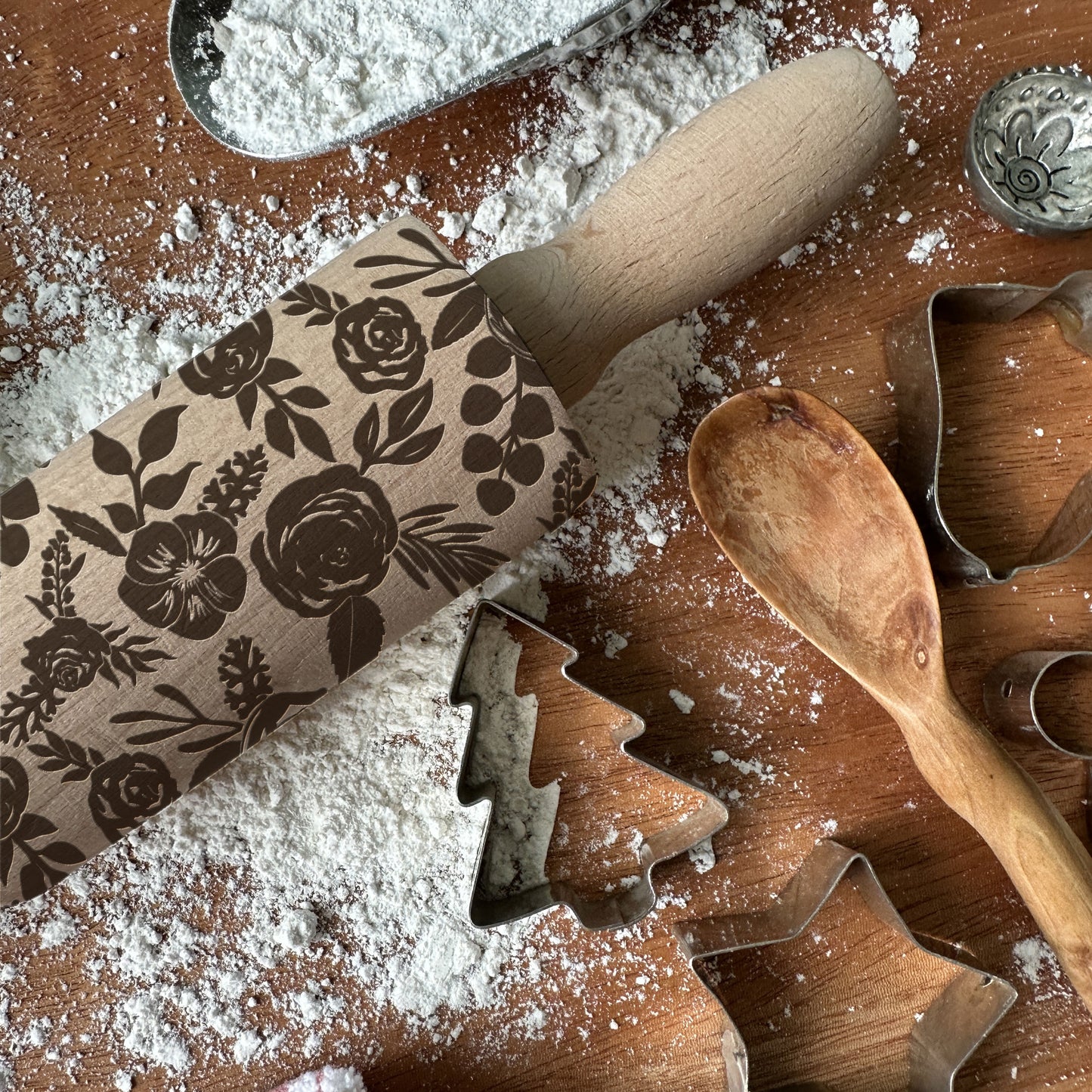 Baking tools including a floral-patterned rolling pin, wooden spoon, and metal cookie cutters on a flour-dusted surface.