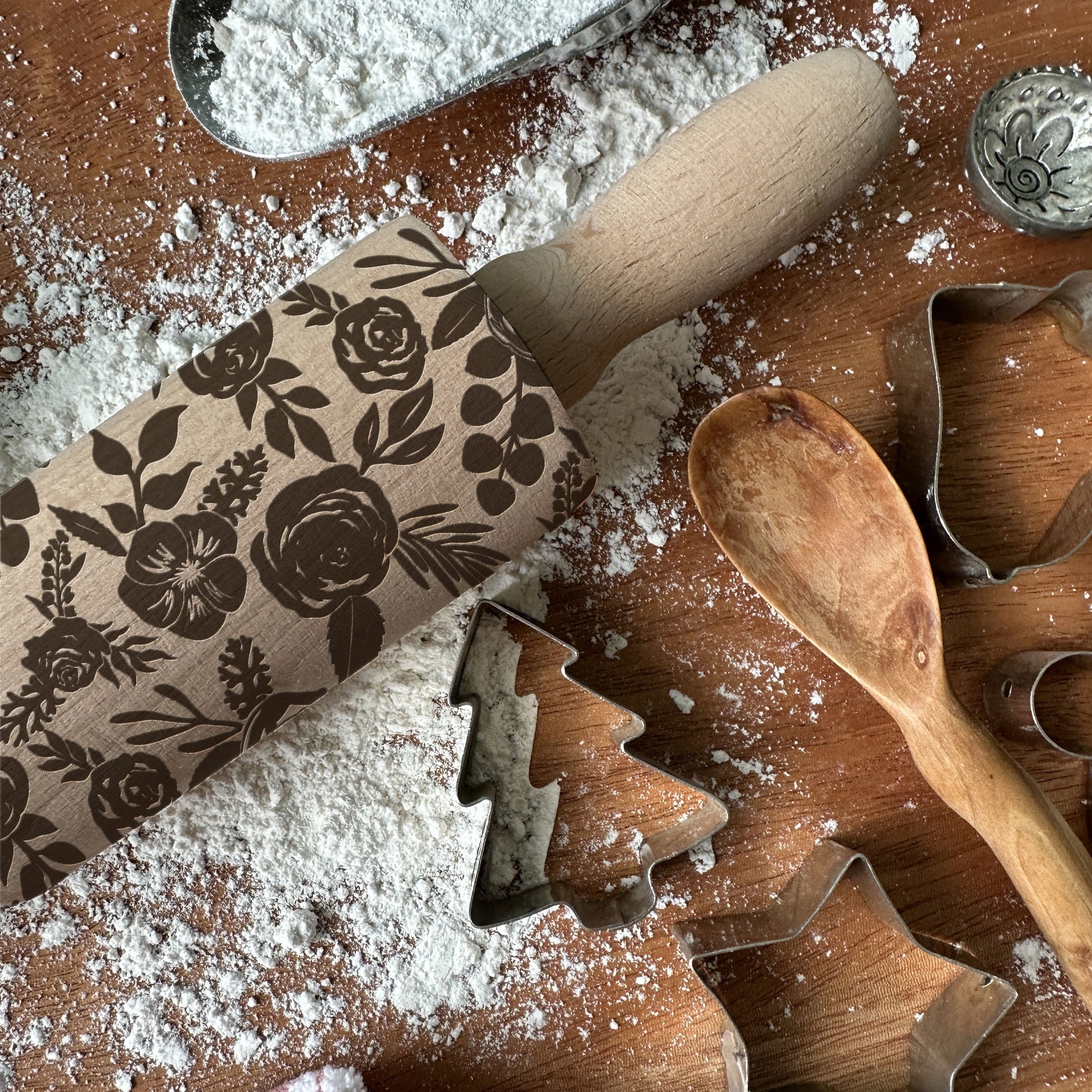 Baking tools including a floral-patterned rolling pin, wooden spoon, and metal cookie cutters on a flour-dusted surface.