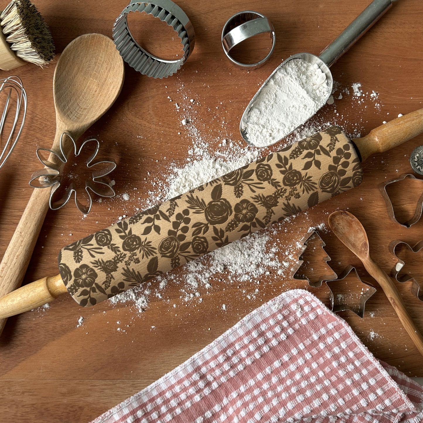 Baking tools including a floral-patterned rolling pin, wooden spoon, and cookie cutters on a wooden surface with flour.