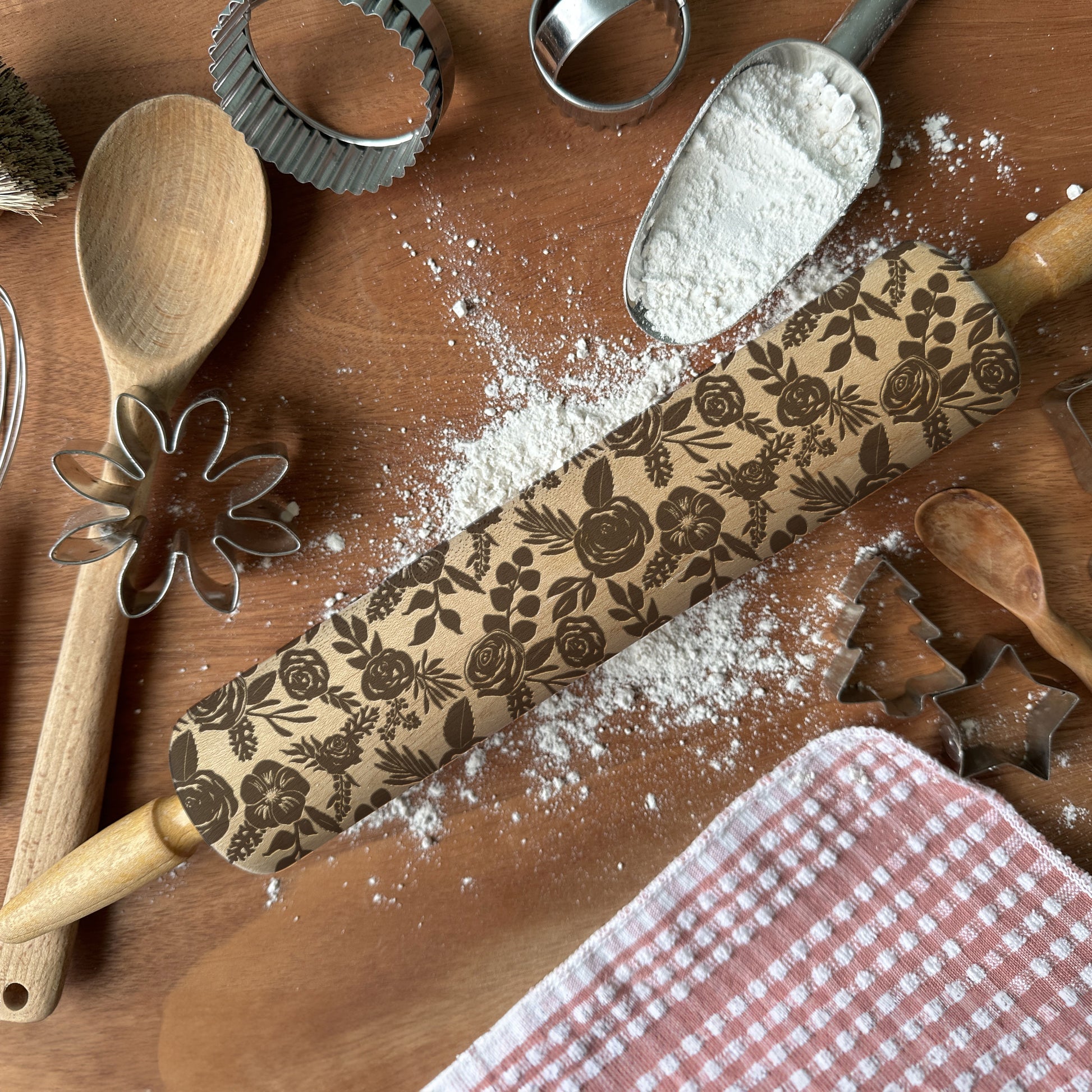 Baking tools including a rolling pin with floral patterns, wooden spoon, flour sifter, and cookie cutters on a wooden surface.
