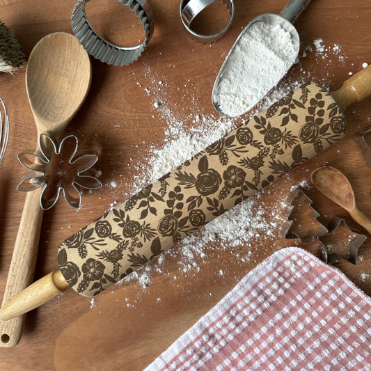 Baking tools including a rolling pin with floral patterns, wooden spoon, flour sifter, and cookie cutters on a wooden surface.