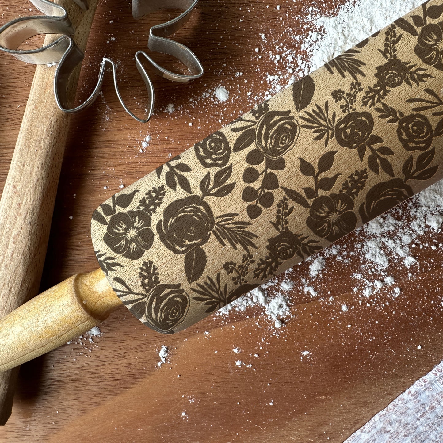Floral-patterned rolling pin on a wooden surface with flour and cookie cutters.