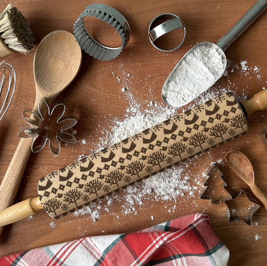 Baking tools including a rolling pin with embossed designs, wooden spoon, whisk, and cookie cutters on a wooden surface with flour.
