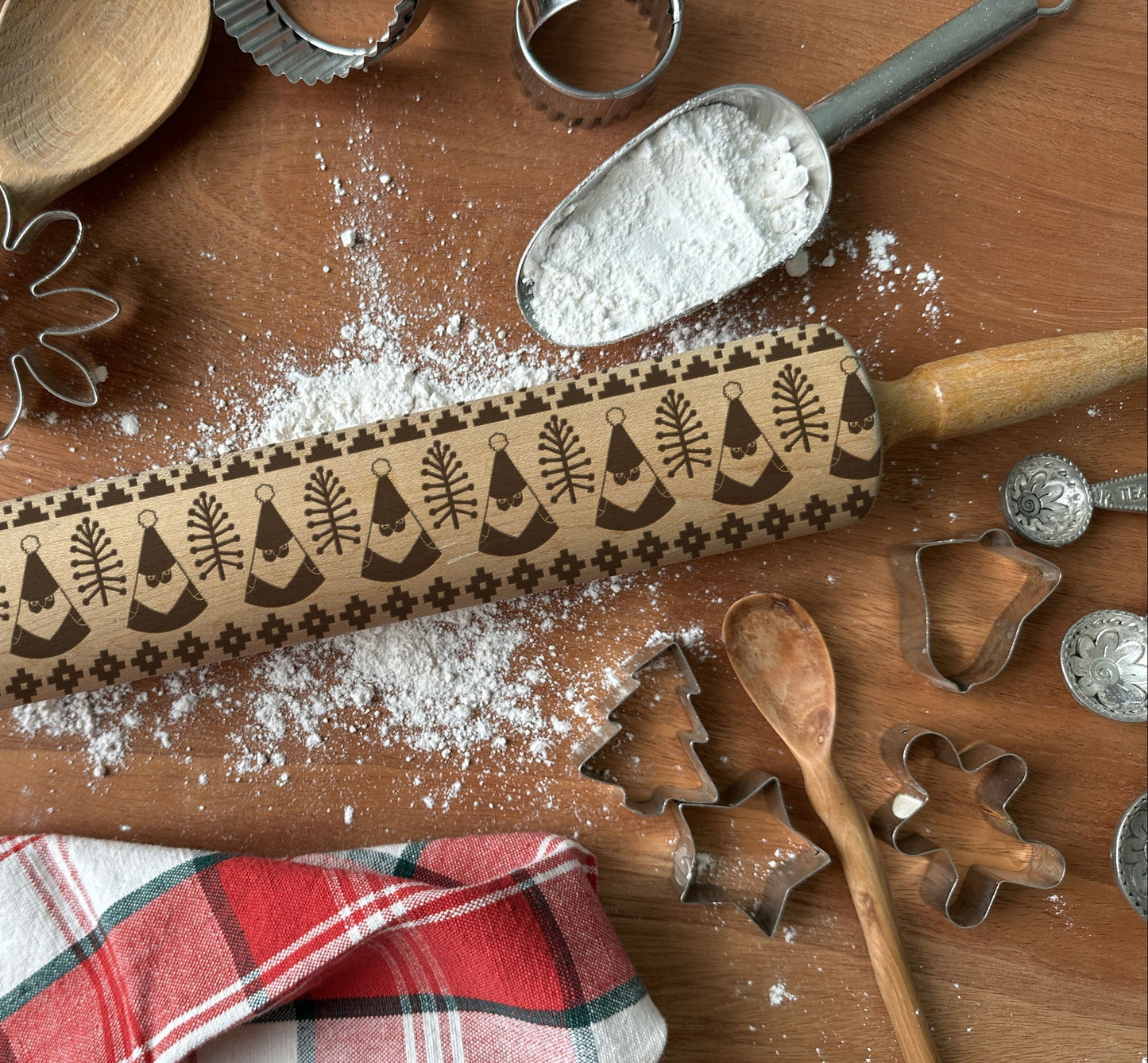 Baking tools including a rolling pin with Christmas designs, cookie cutters, and flour on a wooden surface.